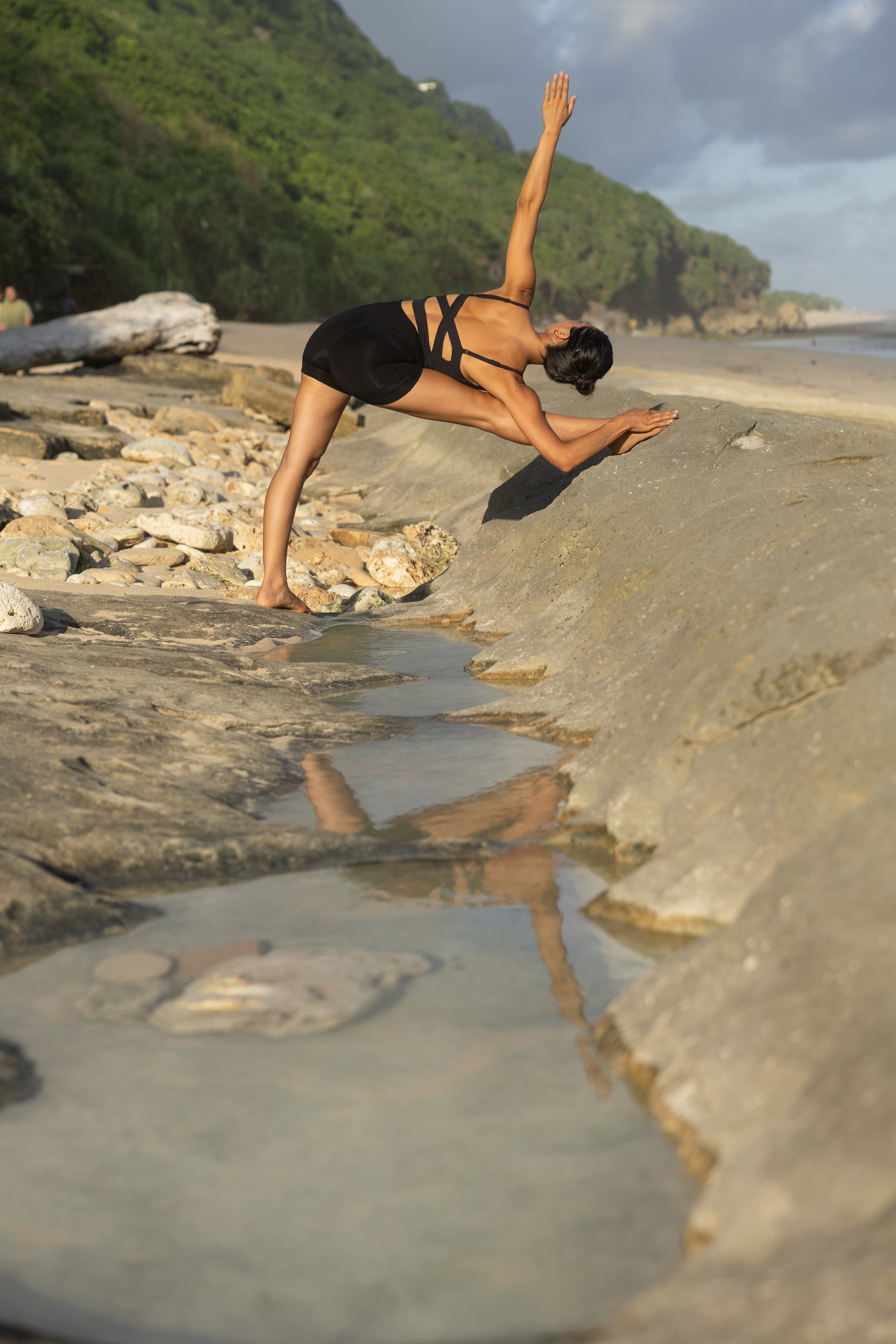 Person practicing yoga on a rocky beach with a scenic background. Women wearing a unique back sports bra.