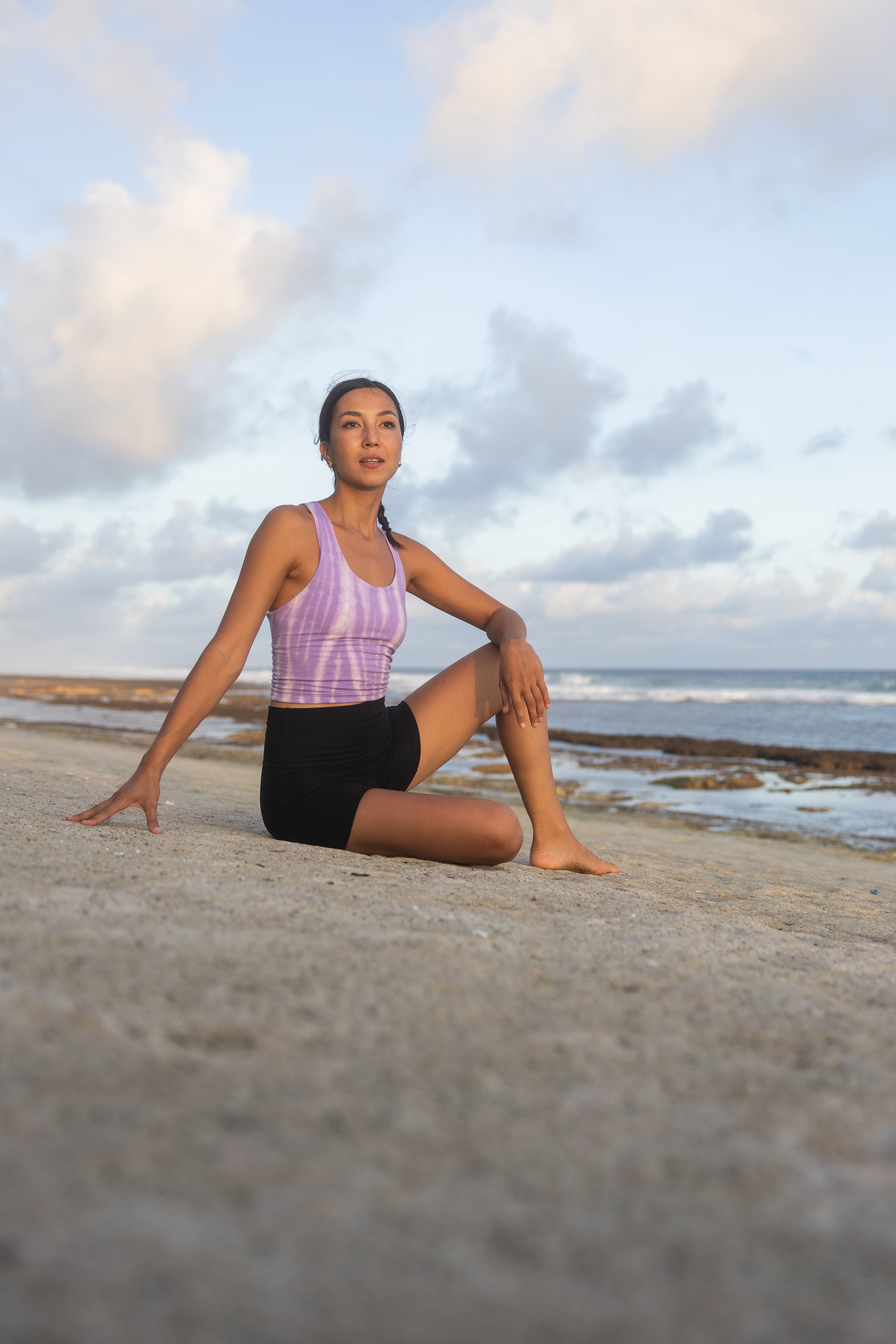 Woman sitting on the beach with a purple top and black shorts, looking towards the ocean.